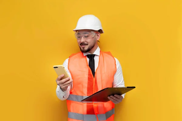 asian civil engineer in uniform uses smartphone on yellow isolated background, male Korean in hard hat and work vest is typing on phone, architect worker