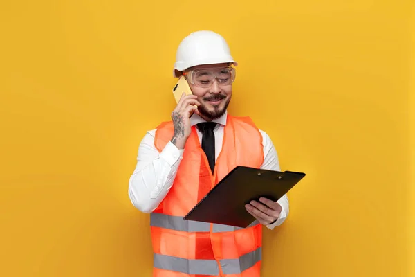 asian builder engineer in uniform talking on the phone on yellow isolated background, male korean in hard hat and work vest communicates on smartphone, architect worker