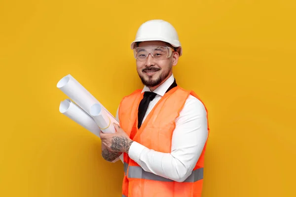 asian builder engineer in uniform holding papers with blueprints on yellow isolated background, male korean in hardhat and work vest, worker architect