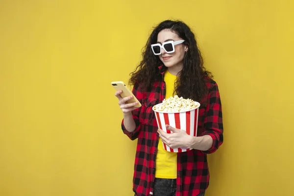 Young Curly Girl Glasses Uses Smartphone Holds Popcorn Yellow Background — Stock Photo, Image
