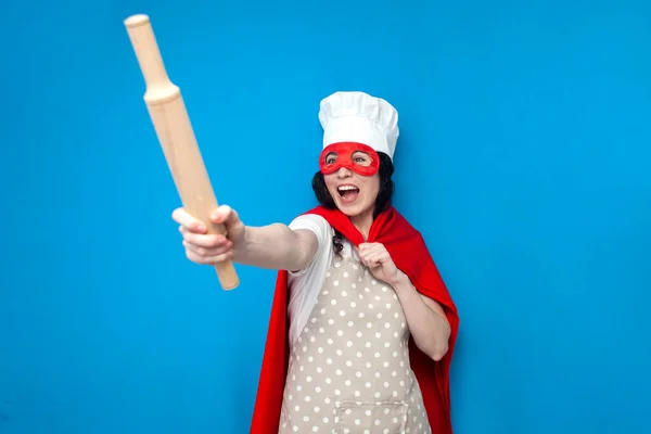 身着超人服装的女厨师拿着蓝色背景的厨房用品 戴着超级英雄面具的女家庭主妇向前看了看 — 图库照片