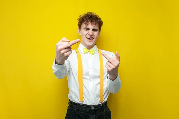 young angry guy in festive outfit shows the middle finger on yellow isolated background, nerd student in shirt with bow tie and suspenders shows fuck