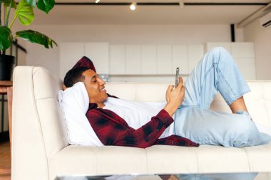 Genç Afrikalı Amerikalı adam rahat bir kanepeye uzanıyor ve akıllı telefon kullanıyor, adam evde telefonu tutuyor ve gülümsüyor, adam dinleniyor ve elinde cep telefonu tutuyor.
