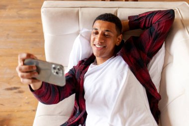 Genç Afrikalı Amerikalı adam rahat bir kanepeye uzanıyor ve akıllı telefonuyla selfie çekiyor, telefonlu adam evdeki video bağlantısıyla iletişim kuruyor, adam kendini kameraya çekiyor.