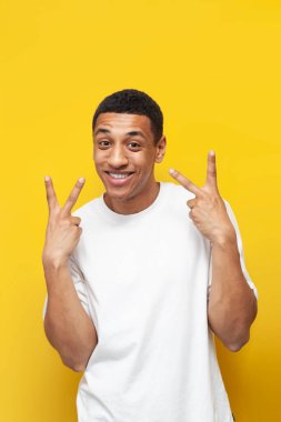 Beyaz tişörtlü genç Afro-Amerikan adam ellerini sarı izole bir arka planda barış jesti gösteriyor, adam iki parmak gösteriyor, dikey çerçeve