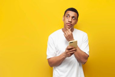 Beyaz tişörtlü genç dalgın Afrikalı Amerikalı adam, sarı izole edilmiş arka planda akıllı telefon ve rüyalar kullanıyor, adam telefonuyla fotokopi alanına bakıyor ve düşünüyor.