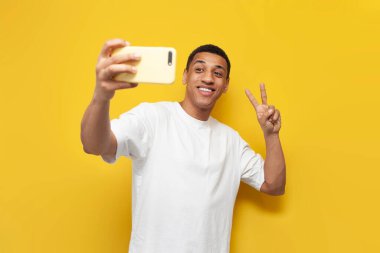 Beyaz tişörtlü genç Afrikalı Amerikalı adam selfie çekiyor ve sarı izole edilmiş arka planda barış jesti gösteriyor, erkek blogcu internet üzerinden video bağlantısı üzerinden iletişim kuruyor