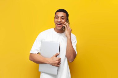 Beyaz tişörtlü genç Afro-Amerikan adam dizüstü bilgisayarı tutuyor ve telefonda sarı izole edilmiş arka planda konuşuyor, modern aletleri olan adam akıllı telefondan iletişim kuruyor.