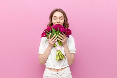 Beyaz şenlik elbiseli genç ve çekici bir kadın pembe lale buketi tutuyor ve pembe izole arka plandaki aromayı kokluyor. Çiçekli kız, bahar ve kadın bayramı kavramı.
