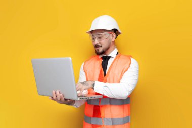 Üniformalı Asyalı ustabaşı, sarı izole edilmiş arka planda dizüstü bilgisayar kullanıyor. Koreli inşaat mühendisi, sabit şapka ve gözlük takıyor ve bilgisayar kullanıyor. İnternette çalışıyor.