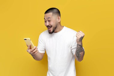 Beyaz tişörtlü yetişkin Asyalı adam akıllı telefon tutuyor ve sarı izole edilmiş arka planda zaferle seviniyor. Şanslı Koreli adam telefona bakıyor ve zaferi kutluyor.