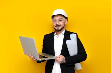 Siyah takım elbiseli Asyalı ustabaşı, sarı izole edilmiş arka planda dizüstü bilgisayar kullanıyor. Koreli inşaat mühendisi, sabit şapka ve gözlük takıyor ve bilgisayar kullanıyor. İnternette çalışıyor.