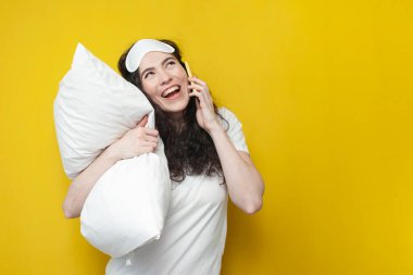 Uyku maskeli tatlı genç kız yumuşak yastığı tutuyor ve yatmadan önce telefonla konuşuyor, pijamalı kadın sarı arka planda cep telefonuyla iletişim kuruyor.