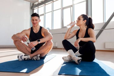 Spor giyimli güzel bir çift sabahları spor salonunda yoga minderlerine oturur ve gülümserler. Spor salonunda antrenman yapan ve su içen atletik erkek ve kadın.