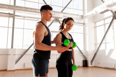 beautiful fitness girl in sportswear trains with a trainer in bright gym, man trainer helps woman lift dumbbells at fitness training