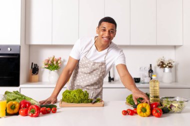 Genç Afrikalı Amerikalı önlüklü adam sebzeli vejetaryen salatası hazırlıyor ve modern mutfakta sebzeler ve yeşillikler, ev aşçısı adam yeşillikleri kesiyor.