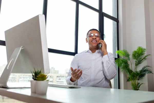 office worker at table in the office sits near computer and talks on the phone, african american business man in the office communicates by cellular communication