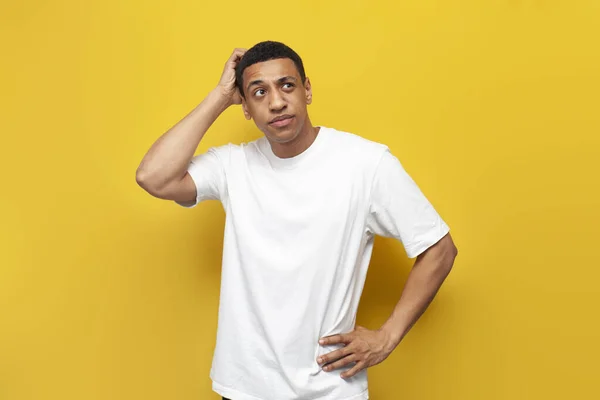 young puzzled african american guy in white t-shirt sketches his head with his hand on yellow isolated background, confused man thinks over problem