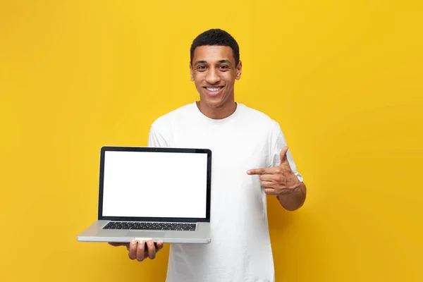 Stock image young african american guy in white t-shirt shows blank laptop screen on yellow isolated background, man advertises computer