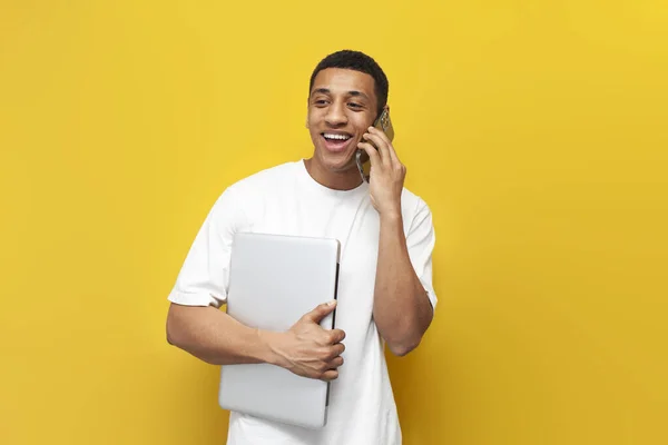 stock image young african american guy in white t-shirt holds laptop and talks on the phone on yellow isolated background, man with modern gadgets communicates on smartphone