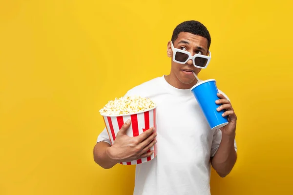 young african american guy in 3d glasses with popcorn and soda looks to the side at copy space on yellow isolated background, male cinema viewer dreams and thinks