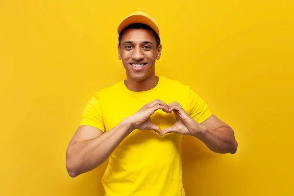 young african american delivery man in yellow uniform showing heart with hands on yellow background, service service worker showing love gesture