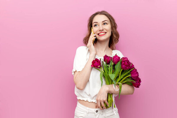 young woman in festive clothes holding bouquet of pink tulips and talking on the phone on isolated background, girl with flowers communicates by cellular, concept of spring and women's holiday