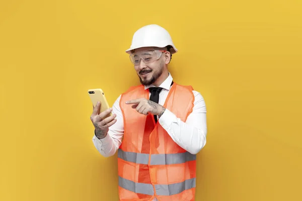 stock image asian foreman in uniform chooses online on smartphone on yellow isolated background, korean civil engineer in hard hat and glasses clicks on the phone