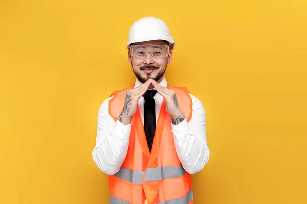 asian foreman in uniform shows house with his hands on yellow isolated background, korean civil engineer in formal wear shows roof gesture, concept of comfort and protection