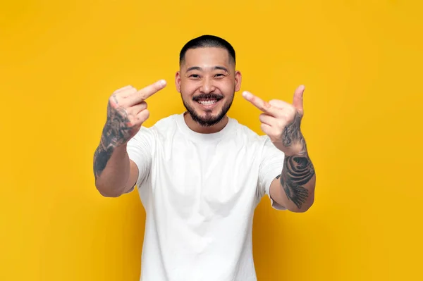 adult rude tattooed asian man in white t-shirt showing middle finger on yellow isolated background, unshaven angry korean guy showing fuck gesture
