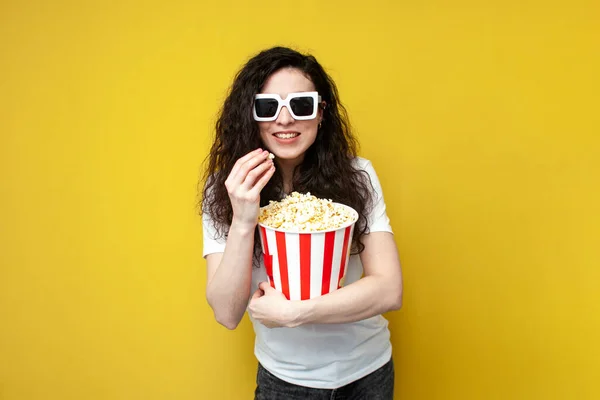 若いです巻きガールビューアで3Dメガネと白のTシャツを保持し 黄色の背景にポップコーンを食べ ブルネットの女性の腕時計映画 — ストック写真