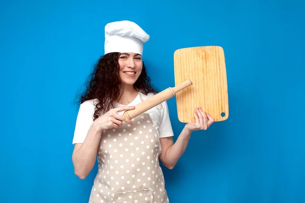 身着制服的女厨师在蓝色背景的复制空间展示 戴着围裙和帽子的女家庭妇女拿着彩色背景的厨房用品 — 图库照片