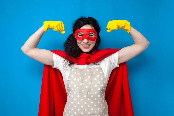 young girl cleaner in gloves and superman costume shows strength on blue background, woman housewife in superhero mask shows biceps
