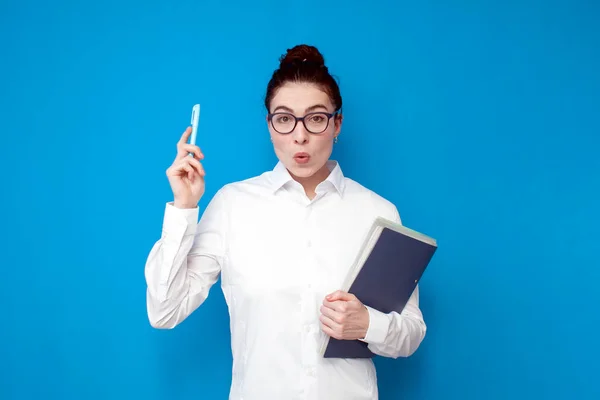 young shocked girl office worker in white shirt and glasses shows surprise with her hand up on blue background, surprised female teacher with documents has idea