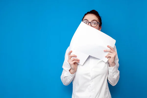 frightened woman manager covers her face with documents on blue background, woman office worker is shy and hides behind papers