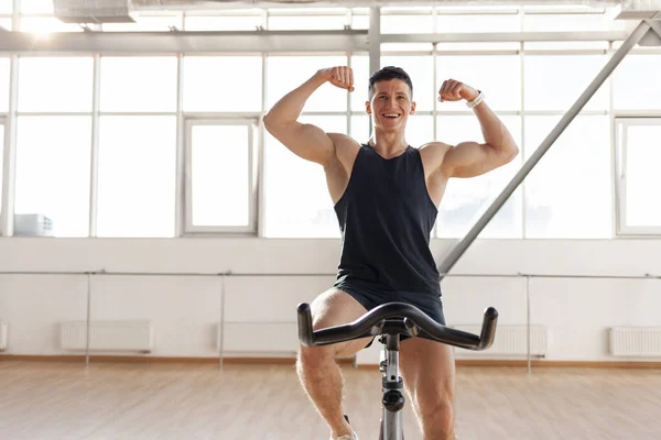 athletic male cyclist trains on exercise bike in bright room in the morning, guy in sportswear sits on simulator in fitness room and shows muscular arms
