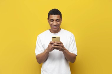 Beyaz tişörtlü genç Afro-Amerikan adam akıllı telefon kullanıyor ve sarı izole edilmiş arka planda mesaj yazıyor. Adam telefonu tutuyor ve gülümsüyor.