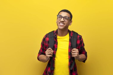 Sırt çantalı Afro-Amerikan öğrenci, sarı izole edilmiş arka plan, tişörtlü Latin öğrenci, öğrenim ve eğitim konsepti üzerine kopya alanına bakıyor.