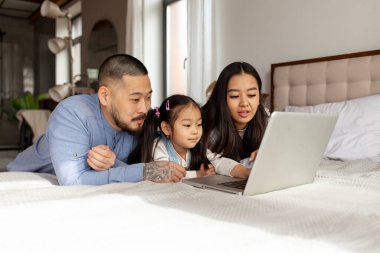 Küçük kızı olan mutlu Asyalı bir aile evde yatar ve dizüstü bilgisayar kullanır, Koreli baba ve kız bilgisayar ekranına bakar.