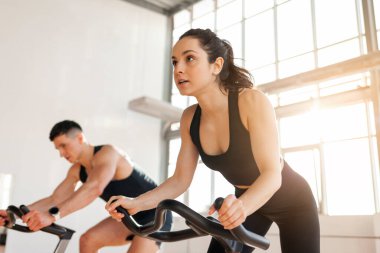 beautiful athletic couple of cyclists train in the fitness room on static bicycle simulator, man trainer and fitness woman go in for sports together on simulators in the morning in bright room