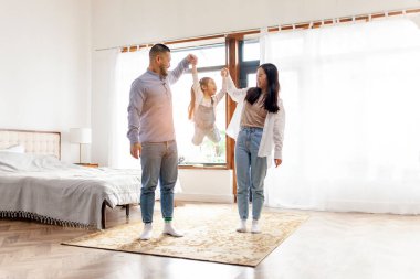 happy Asian family with little daughter stands at home in the bedroom and rejoices, dad and mom raise the hands of cheerful child in their house clipart