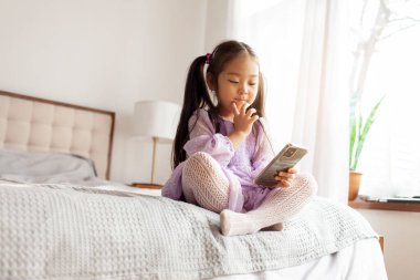 Küçük Asyalı düşünceli kız evde yatakta oturuyor ve akıllı telefon kullanıyor, çocuk telefona bakıyor ve mor elbisesiyle düşünüyor.