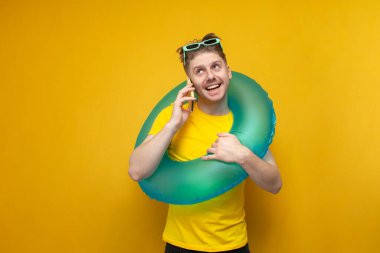 Yaz tatilinde şişme yüzme çemberi olan genç adam sarı arka planda telefonla konuşuyor ve gülümsüyor.