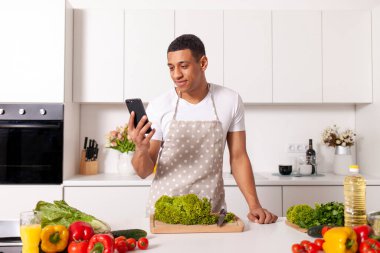 Önlüklü Afro-Amerikan adam modern mutfakta sebzeli ve yeşillikli vejetaryen salatası hazırlıyor ve akıllı telefon kullanıyor, ev aşçısı telefona bakıyor ve internetten malzeme ve ürün seçiyor.