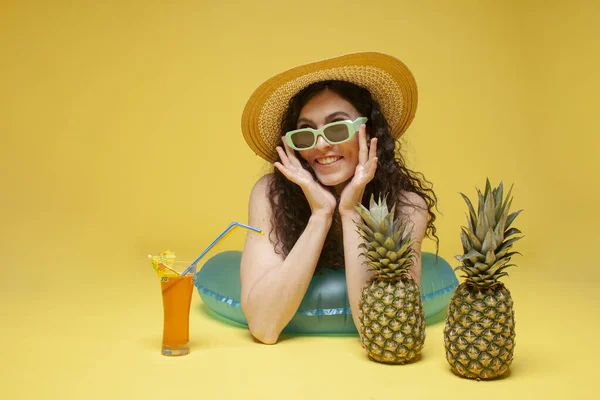 Stock image young girl in a straw hat lies on an inflatable swimming circle with an orange cocktail and pineapples on a yellow background, a woman sunbathes on vacation in the summer