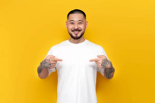 adult tattooed asian man in white t-shirt points to himself on yellow isolated background, korean guy shows his hands on his chest and smiles