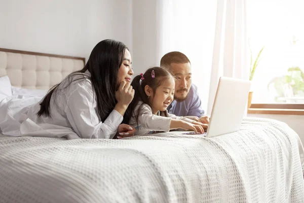 Küçük kızı olan mutlu Asyalı bir aile evde yatar ve dizüstü bilgisayar kullanır, Koreli baba ve kız bilgisayar ekranına bakar.