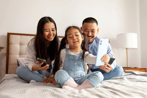 happy asian family with phones sit at home on the bed and communicate via video call, little korean girl with mom and dad takes selfie on smartphone, daughter takes picture of herself with her parents