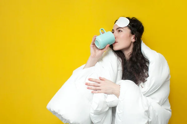 sleepy girl in sleep mask covered with soft blanket drinks coffee on yellow background, the woman woke up in the morning and drinks drink from cup