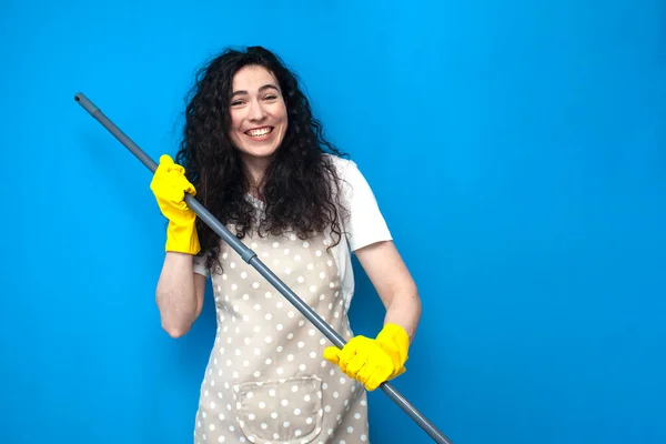 young woman cleaner in apron and gloves for cleaning washes the floor with mop on blue background, girl housewife cleans on colored background, cleaning service worker